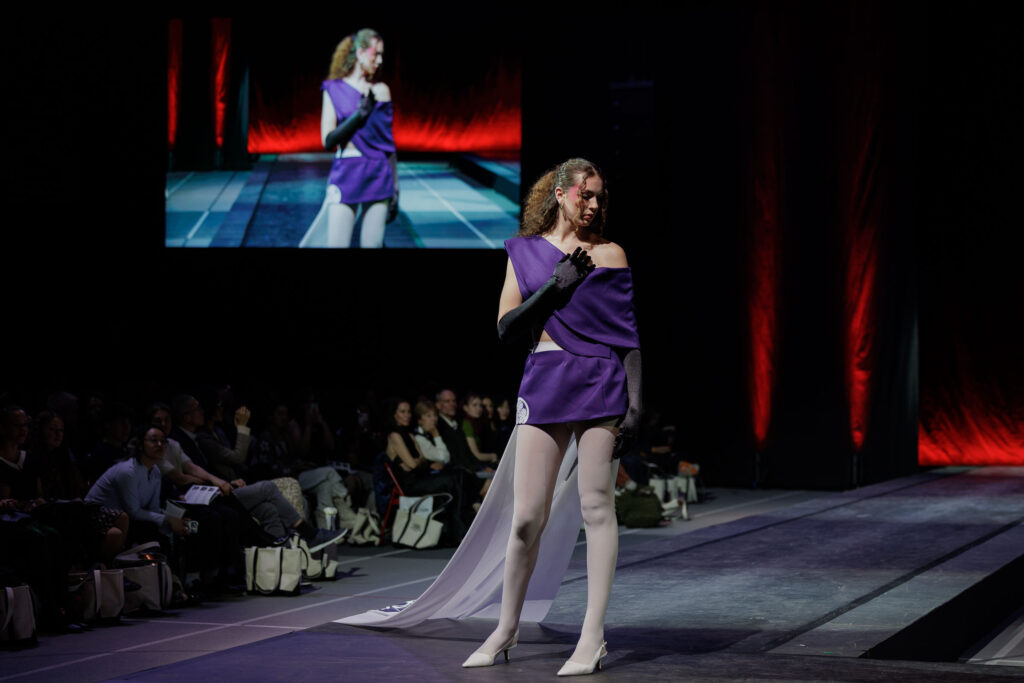 A model poses at the end of the runway wearing a purple top and mini skirt, black gloves, and a white train draping behind her.