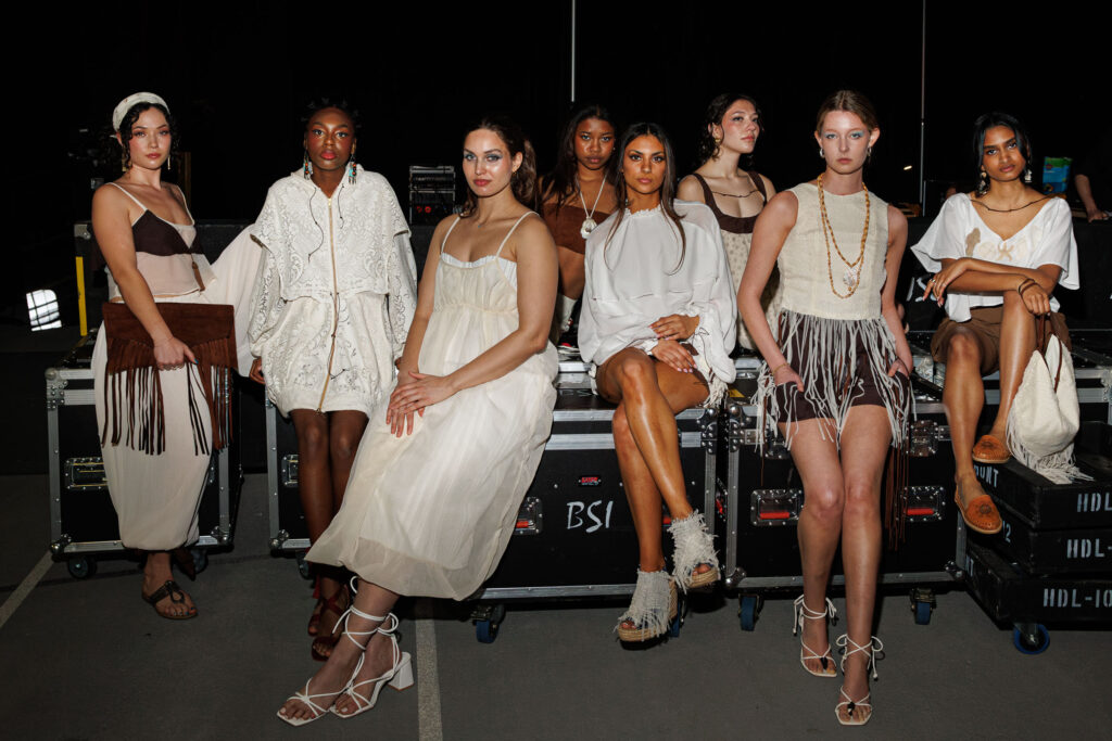 Eight students wearing whites and browns with their hair and makeup done sit backstage.