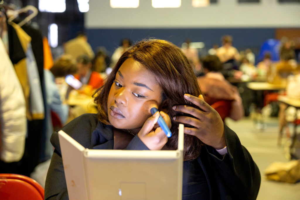 A student applies makeup with a brush while looking in a mirror.