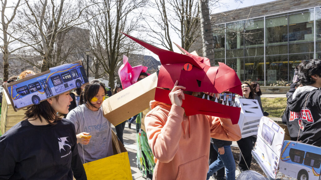 Students in various costumes, including one wearing a red dragon head, walk down the sidewalk together during Dragon Day 2026