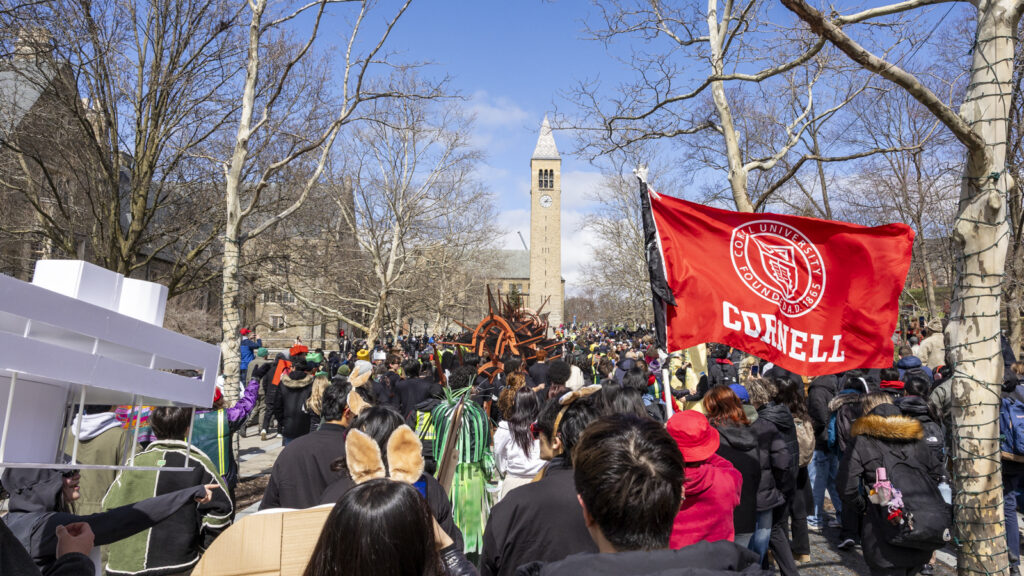 Hundreds of people approach Ho Plaza and McGraw Tower, one waving a red Cornell flag, during the parade at Dragon Day 2026