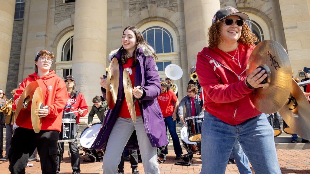 Three students wearing Cornell apparel that are part of a band hold cymbals outside of Goldwin Smith Hall