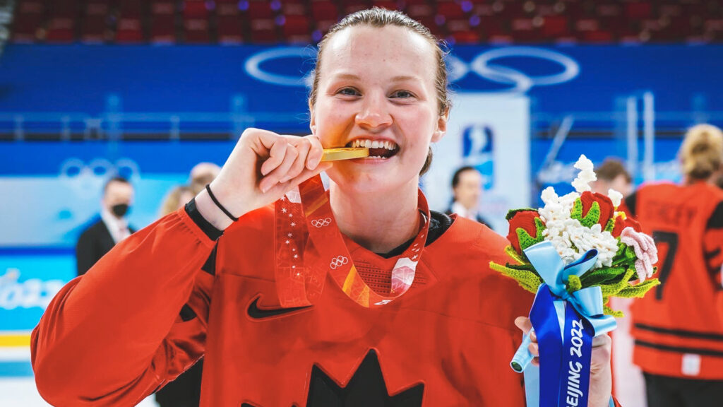 Micah Zandee-Hart smiling and biting her gold medal at the 2022 Olympic Winter Games in Beijing, China.