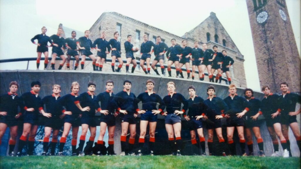 The Cornell Men's Rugby team from 1992 stands in two lines and poses for a photo with their hands on their hips in front of McGraw Tower.