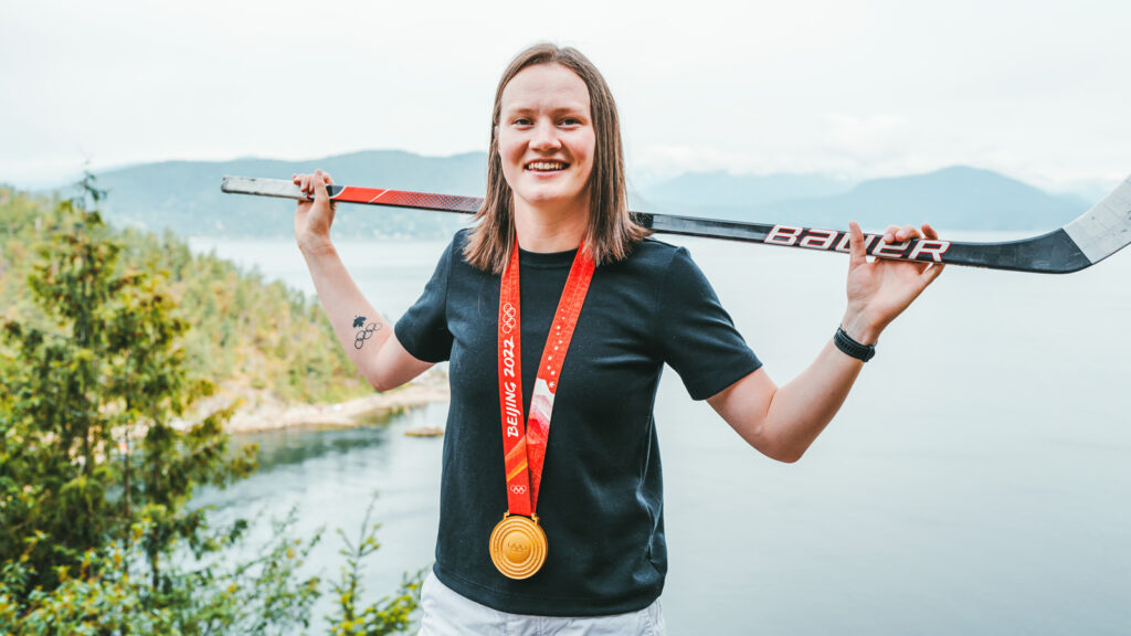 Micah Zandee-Hart with her Olympic gold medal and hockey stick over her shoulders with a lake in the background.