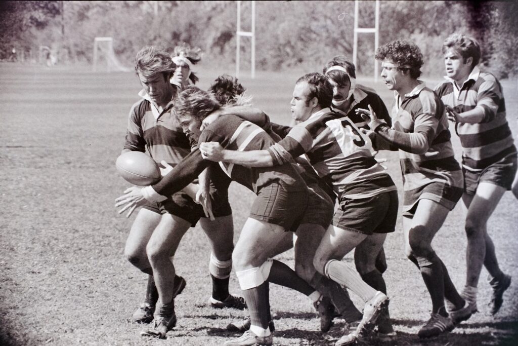A black and white photo of the Cornell Men's Rugby team during a competition running with the ball while other players try to block opponents.