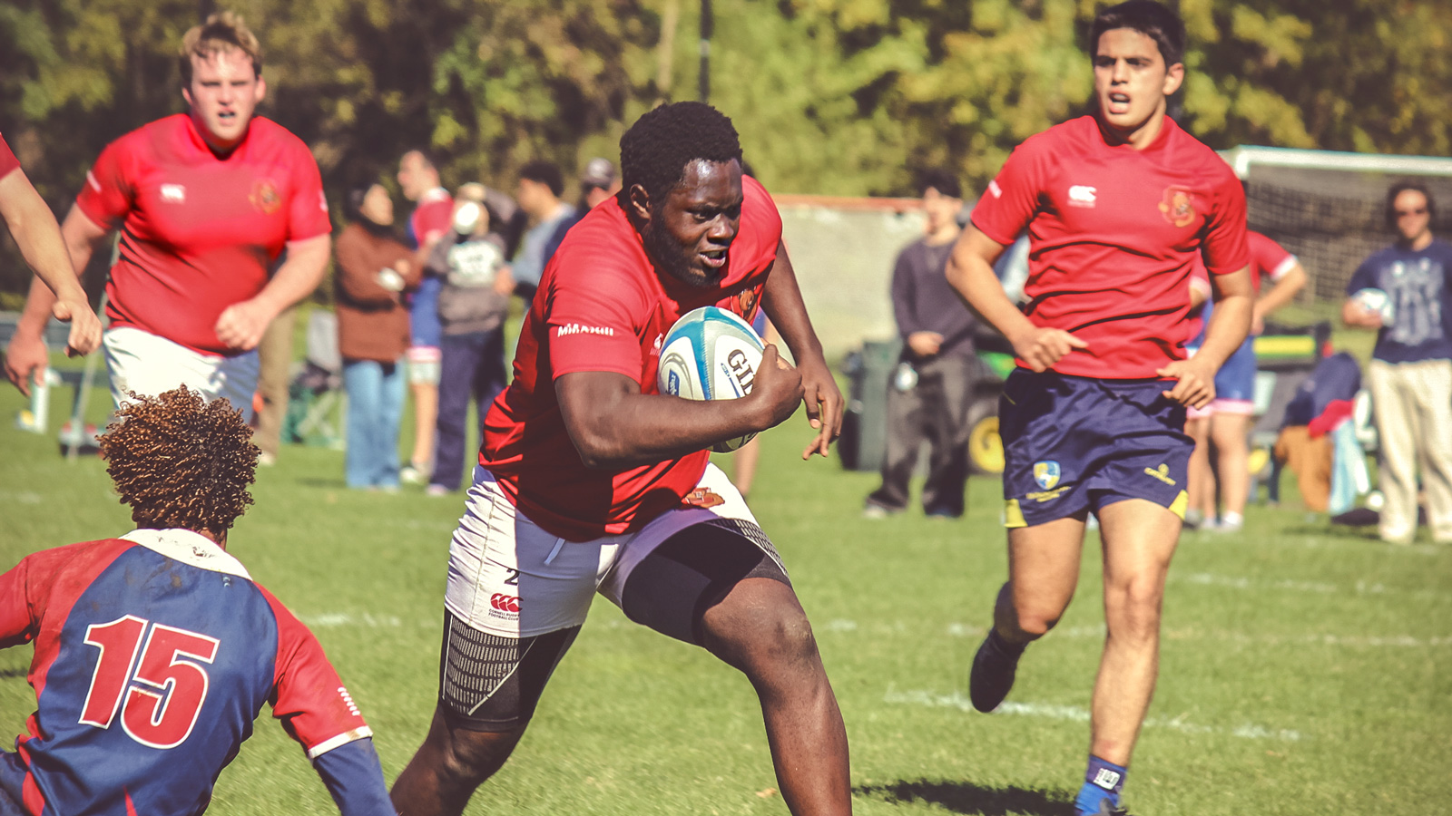 A Cornell men's rugby player runs with the ball and dodges opponents which players in the back cheer for him.