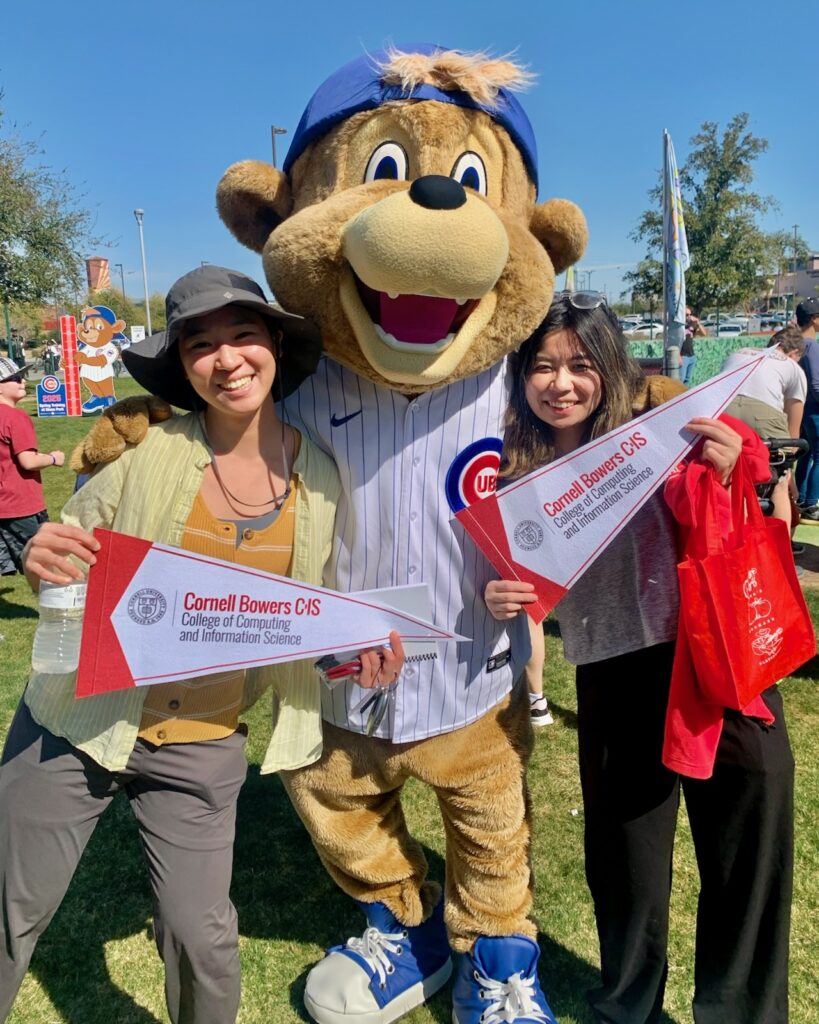 Andrea Wen-Yi Wang and Waki Kamino at a spring training game with Clark, the Chicago Cubs mascot. The students are holding pennants branded with Cornell Bowers CIS.