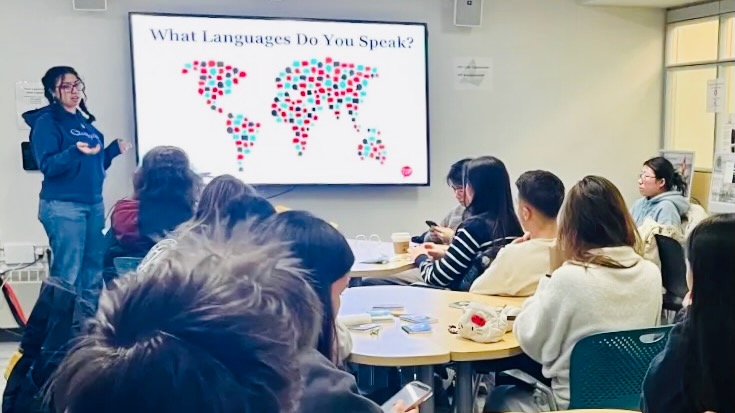 A student presents in front of a smart board to other students sitting around circular desks during an info session.