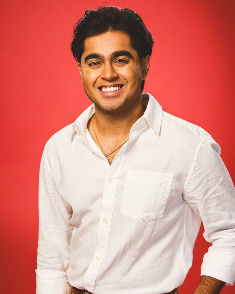 Adi Arora smiles in a white button-down shirt and gold chain in front of a red background.
