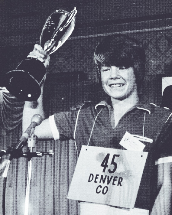 Jacques Bailly holds the National Spelling Bee trophy as a teen in 1980. Around his neck is a sign that says "45 - Denver, CO"