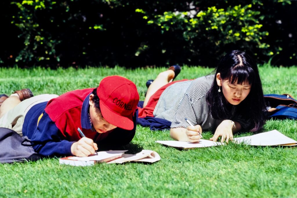 two students study, pens in hand, while lying in the grass on campus in spring 1992