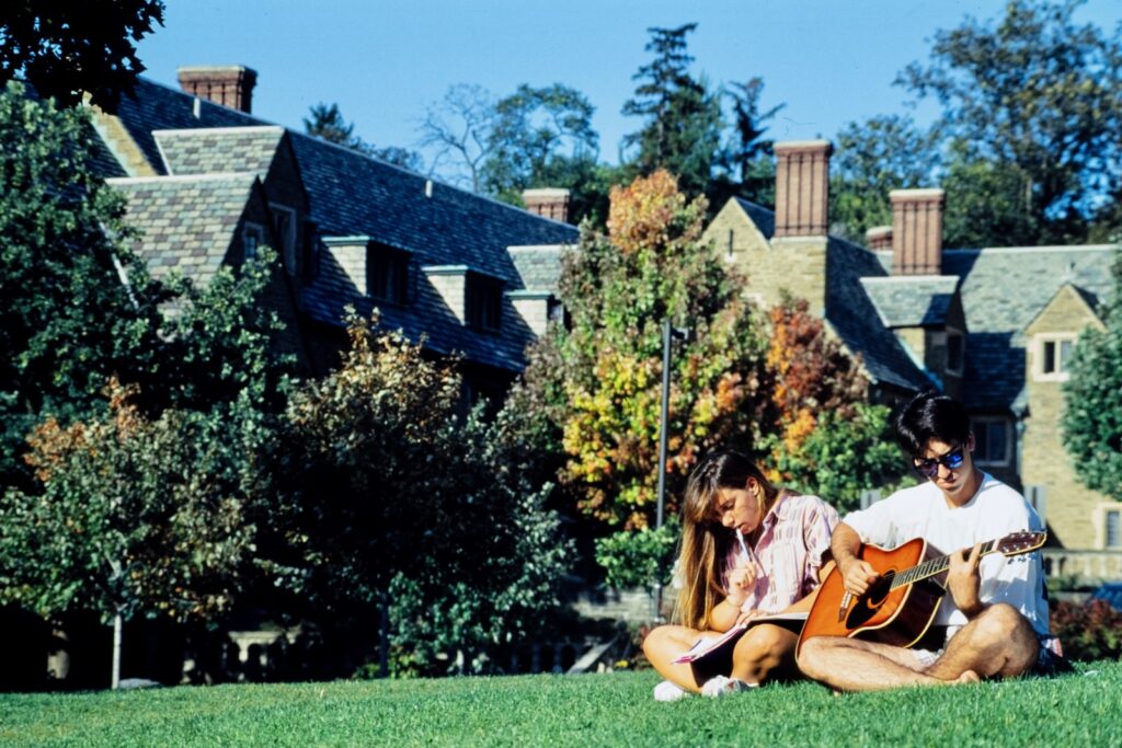 two students sit in the grass on West Campus, one playing guitar, in spring 1992