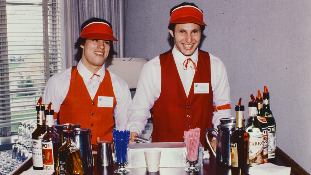 Hotel School students in red vests and visors behind a bar counter at Hotel Ezra Cornell in 1980
