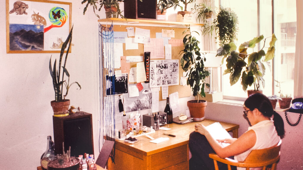 A student sits at her desk studying in the early 1980s, the room decorated with art and plants