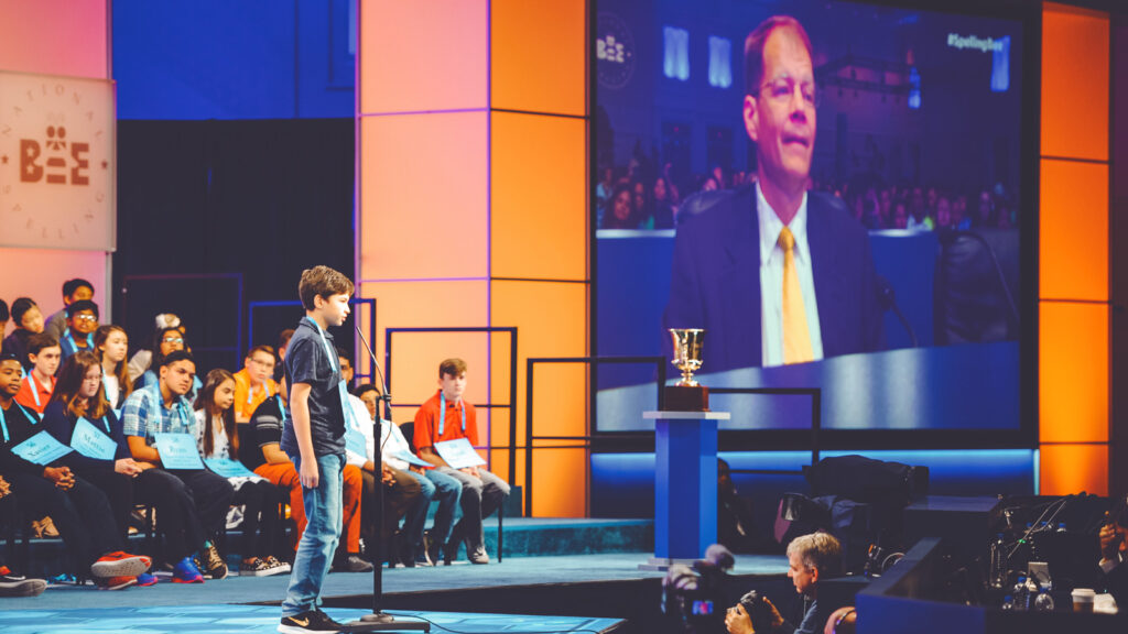 Jacques Bailly is seen on a screen as a boy is at the microphone at the 1980 National Spelling Bee