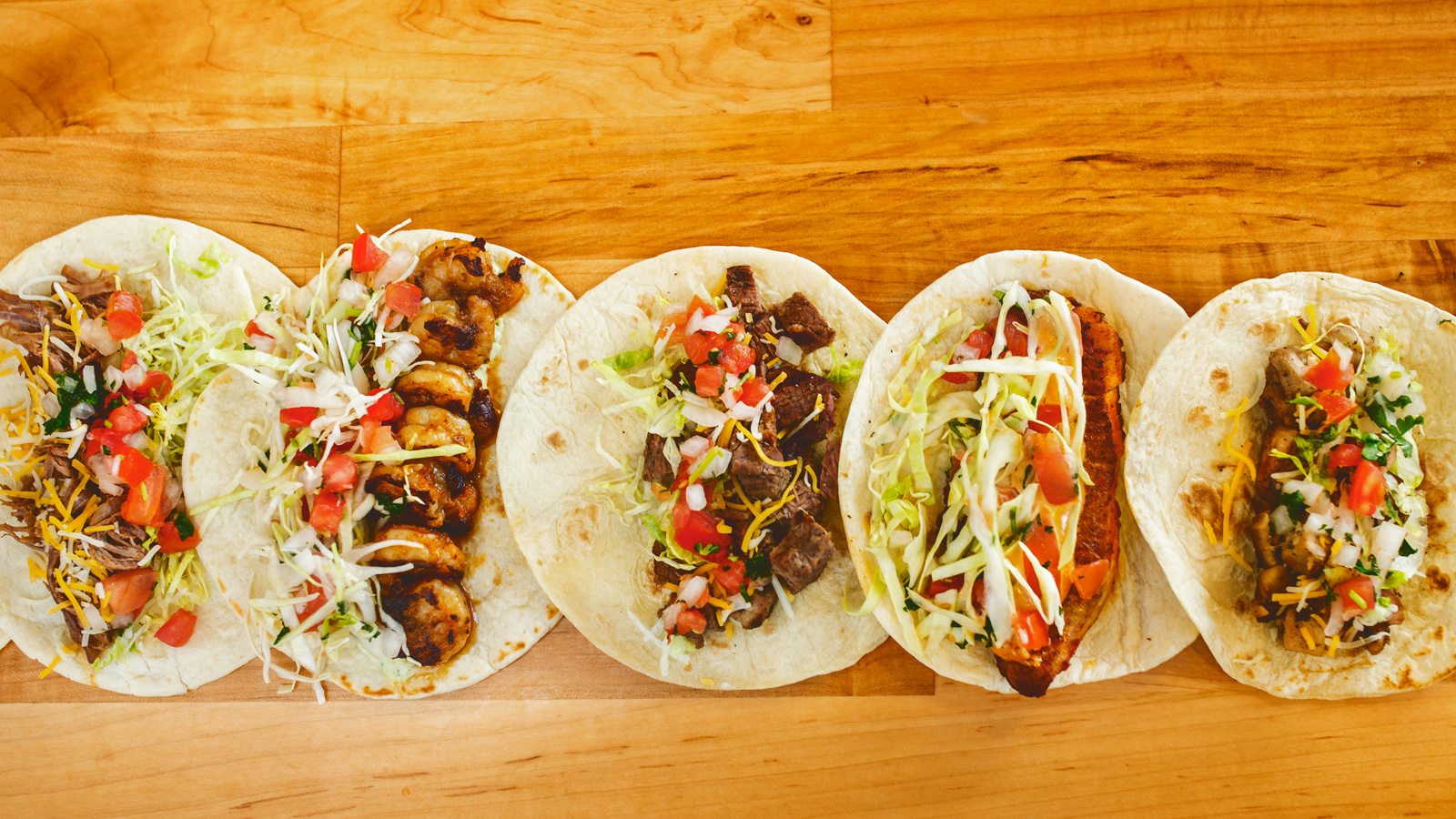 images of several of District Taco's menu items laid out in a row in tortillas on a wooden table surface