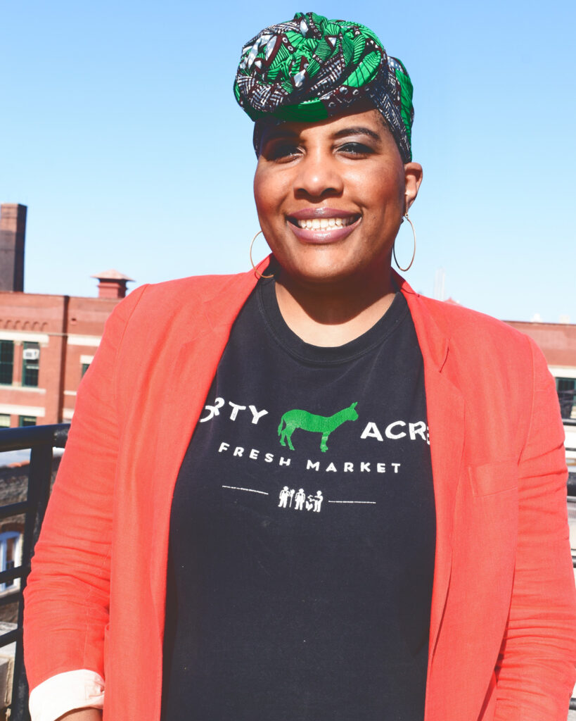 Elizabeth Abunaw smiles while wearing a Forty Acres Fresh Market shirt, a red blazer, and gold hood earrings.