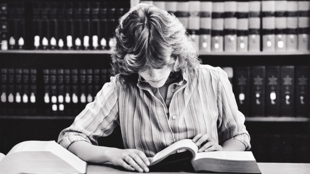 A black and white photo of a student with her head down reading a book in a library