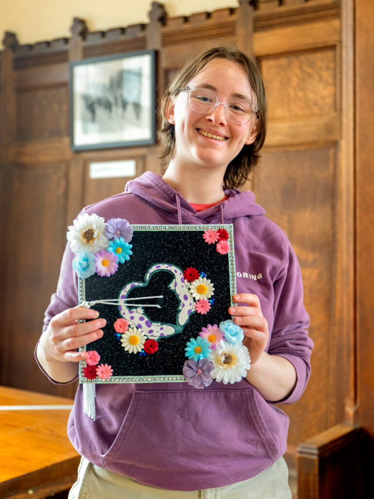 A student holds up her decorated cap, which features a black sparkly base with blue, purple, and pink flowers attached to it.