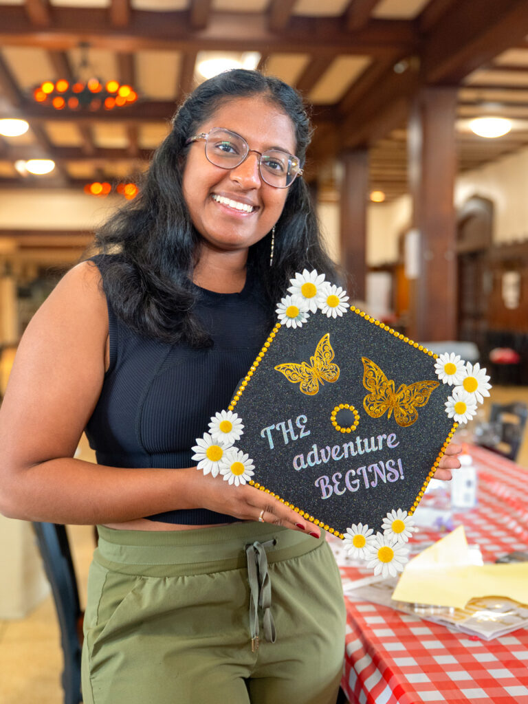 A student holds up her decorated cap, which features gold butterflies, daises, and writing that says "the adventure begins!"