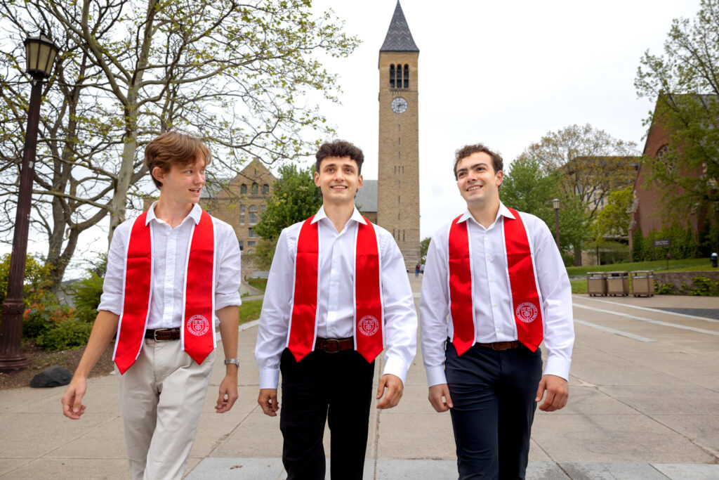 Three students in their red stoles and white button down tops walk together on campus in front of McGraw Tower.