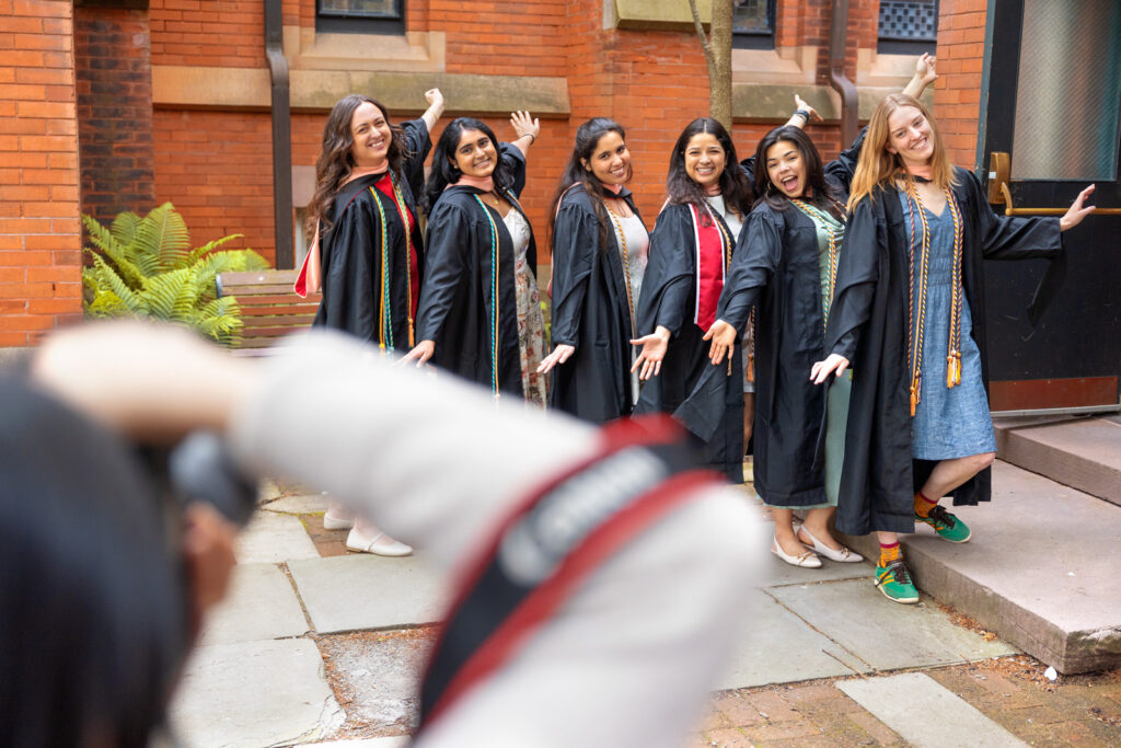 Six students in their black graduation robes, stoles, and cords pose for a picture with their hands in the air.