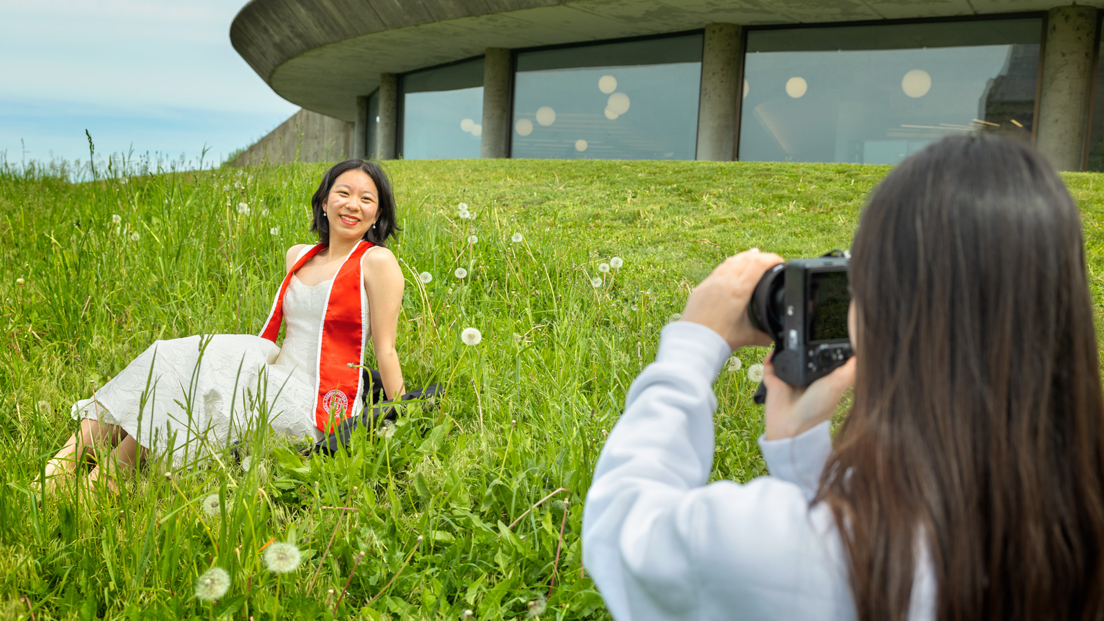 A student in a white dress and red stole sits in the grass and poses for a photo next to Uris Library.