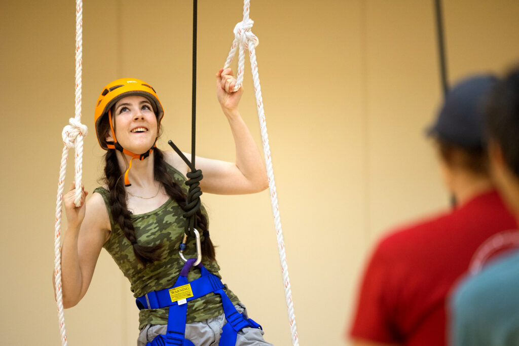 A student wearing a helmet and harness holding two ropes participates in the Senior Days 2026 Indoor Challenge Course in Noyes Gym.