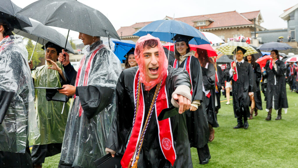 A male grad in the procession wears a red plastic poncho hood and points at the camera
