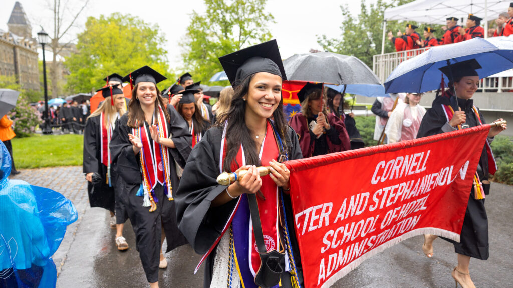 Grads carrying a banner that says Cornell Peter and Stephanie Nolan School of Hotel Administration