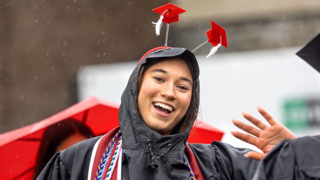 A grad in a rain jacket wearing a pair of novelty ears shaped link small red mortarboards