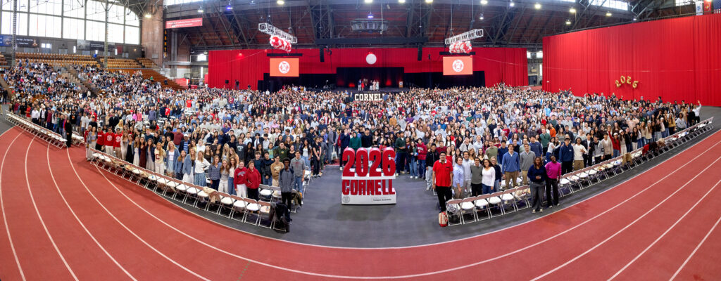Students from the class of 2026 stand together and smile for a photo in Barton Hall during Convocation in front of a sign that says "2026 Cornell."