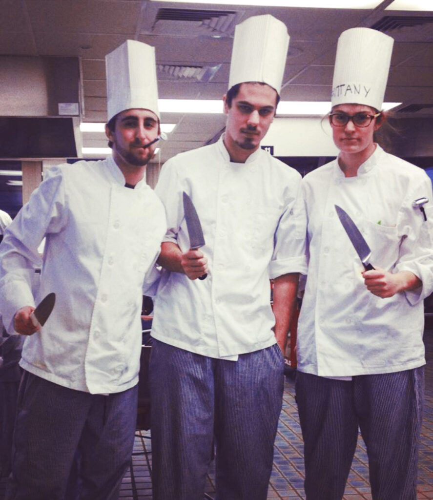 Jake Potashnick and two classmates holding knifes while wearing chef whites during a culinary class.