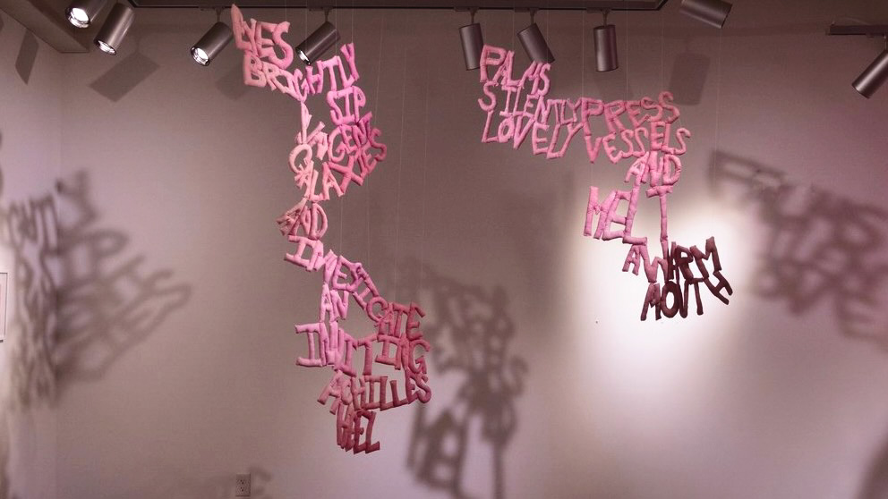Pink fabric words hang from the ceiling while spotlights cast shadows of the work on the white wall behind it.