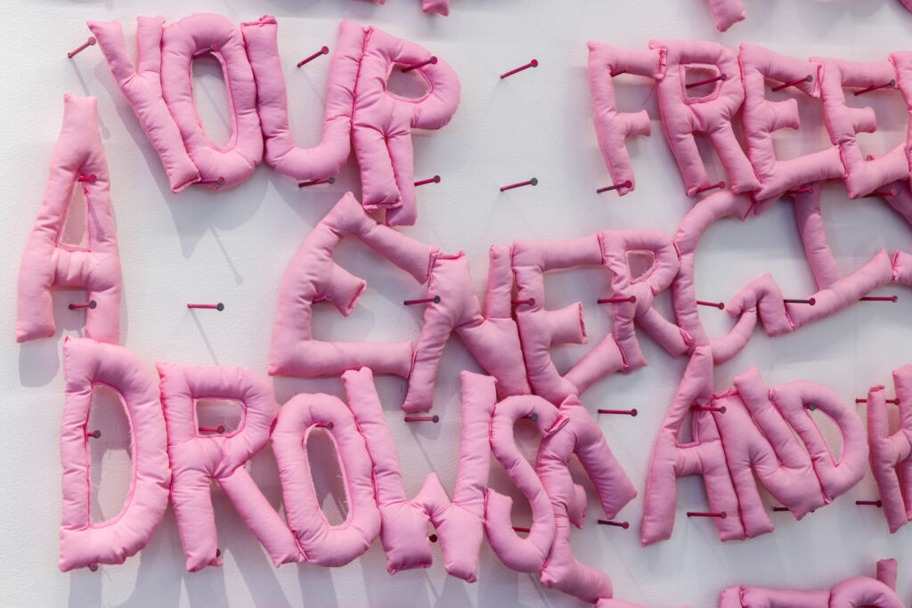 Puffy, pink fabric words are held to a white wall by nails.
