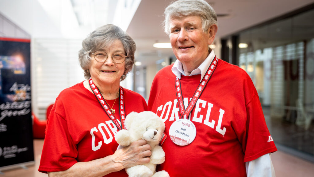 Two alumni wearing red Cornell shirts and Reunion buttons smile for a photo together while one holds a white stuffed bear at Reunion 2026