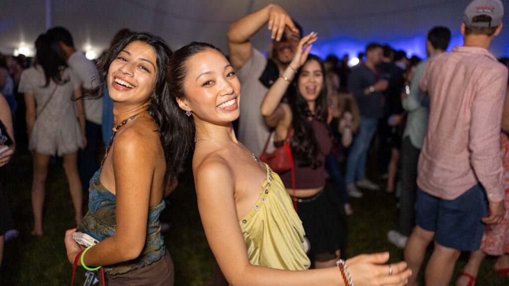 Two girls stand back to back and smile during a tent party while onlookers smile in the background at Reunion 2026