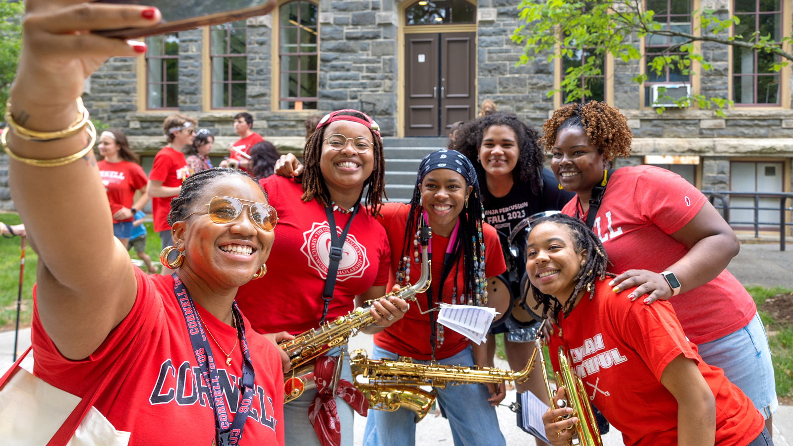 A group of women in red t-shirts, some holding brass instruments, take a selfie on the Arts Quad during Reunion 2026