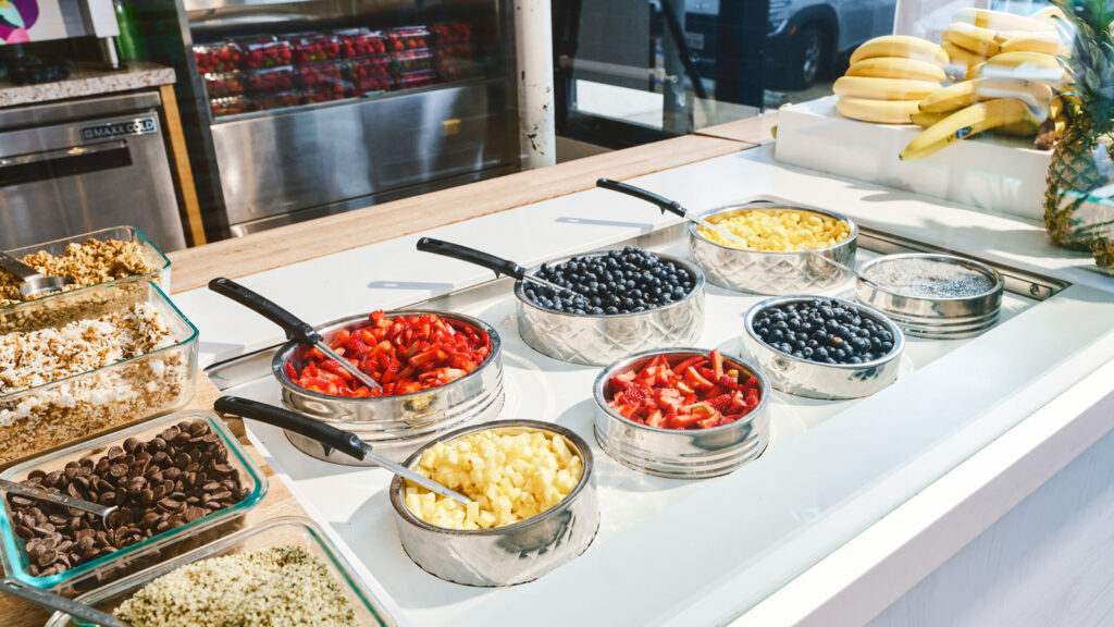 The counter at a SAMBAZON store, featuring containers of strawberries, blueberries, and mangoes as well as a pile of banana and dry toppings including granola.