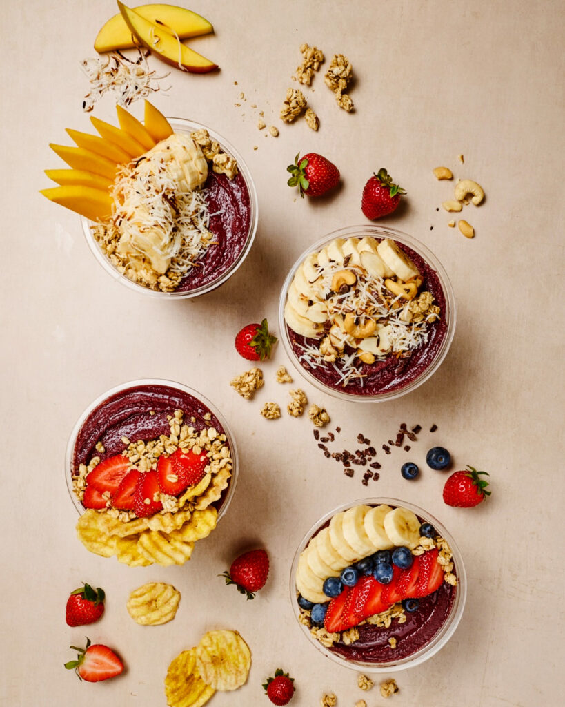 An overhead view of four acai bowls with fruit, granola, coconut, and other toppings on them and around the bowls.