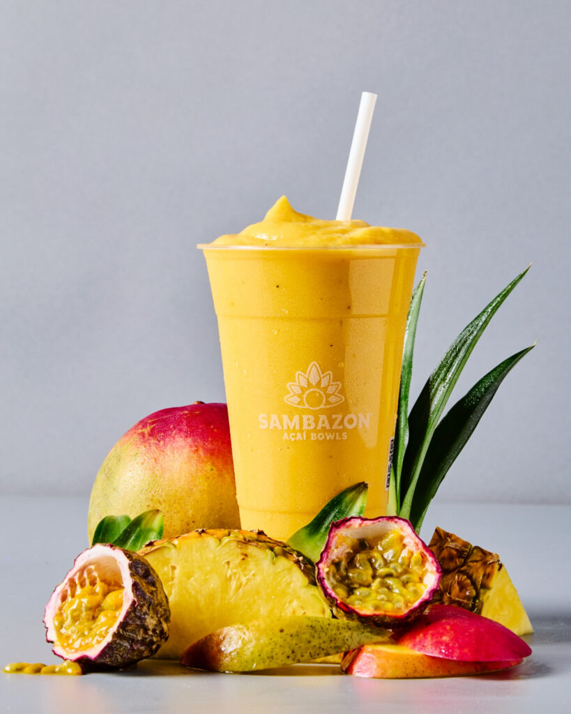 A bright yellow mango smoothie in a SAMBAZON cup with pineapples and mangos around the cup.