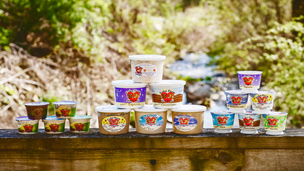 Seventeen containers of Echo Farms pudding in different flavors are stacked on top of each other on a wooden ledge.