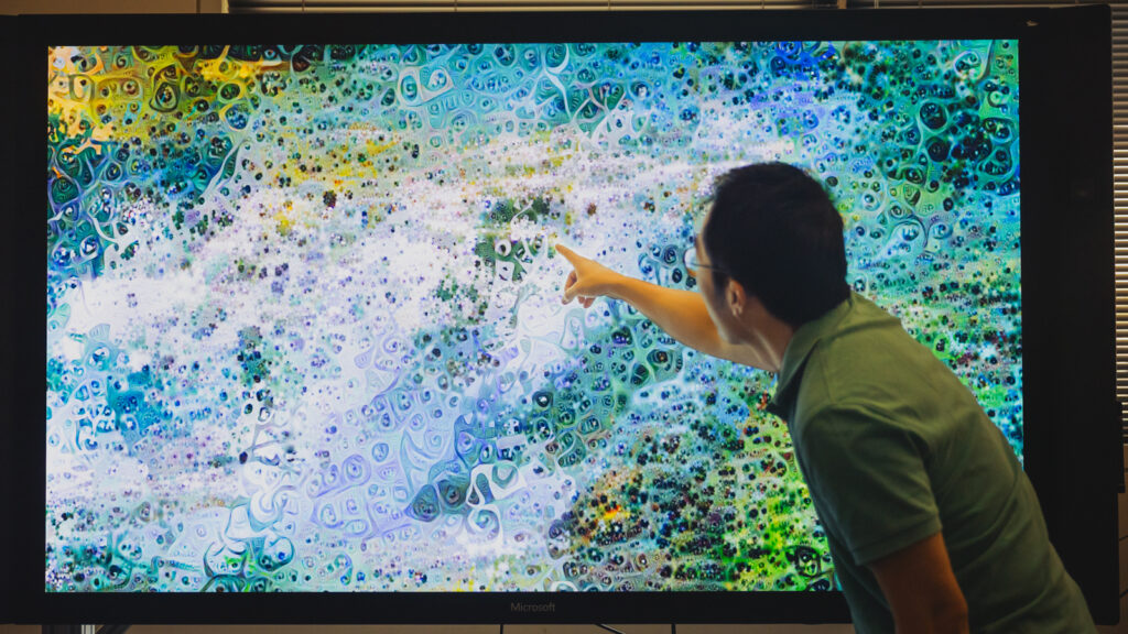 Prof. Alex Kwan points to a piece of artwork on a screen that resembles a biological microscope image, in white, blue, green, and yellow hues