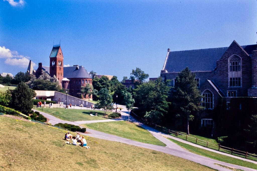 students on the grassy hill between McGraw Tower and Willard Straight Hall, with Barnes Hall and the Cornell Store visible in the background, in a vintage color photo from 1990
