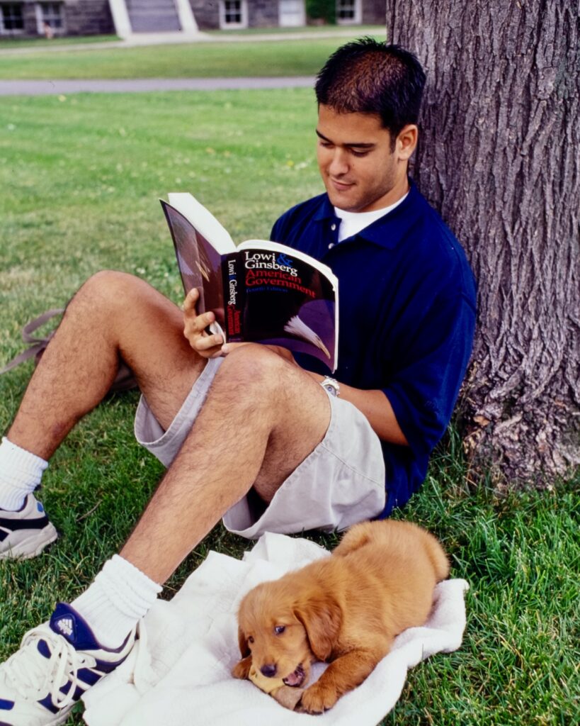a student reads "American Government" outside by a tree, accompanied by a puppy chewing on a bone, in 1997