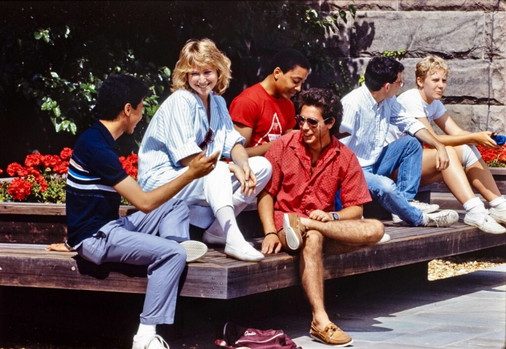 a group of students chat on a wooden bench on campus in a vintage color image from 1986