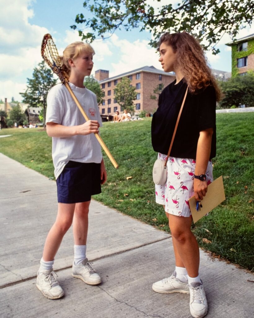 two students, one holding a lacrosse stick, stand on a sidewalk near the U-Halls on West Campus in 1990