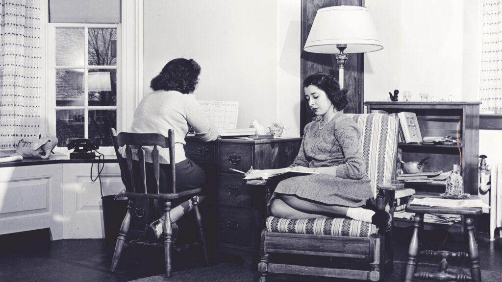 Two female students sit in a dorm room, which has a rotary telephone visible on a shelf
