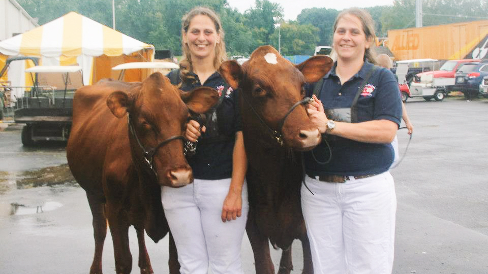 Beth and Courtney Hodge as teens stand with two cows and wear black shirts with the Echo Farms logo on them.